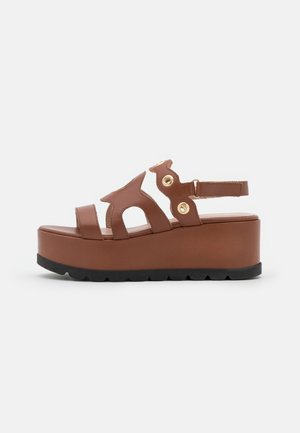 Bruine platformsandaal met dikke zool, twee voorstraps, uitgesneden zijkantontwerp, gouden oogjesdetails en verstelbare enkelriem op witte achtergrond.