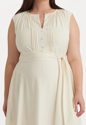 Vrouw draagt een mouwloze crèmekleurige jurk met stoffen knopen, verticale plooien op de borst en een gestrikte tailleband.