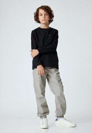 Chemise noire à manches longues avec texture côtelée, pantalon cargo gris avec poches latérales et baskets blanches avec accents bleus.