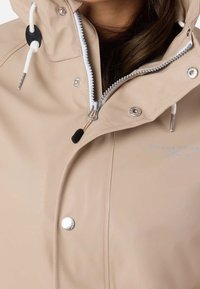 Veste imperméable beige avec fermeture éclair, garnitures argentées, capuche réglable avec cordon de serrage et logo sur le tissu.