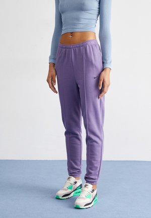 Personne portant un jogging Nike violet, un crop top bleu à manches longues et des baskets blanches Nike avec des accents verts, debout sur un sol bleu.