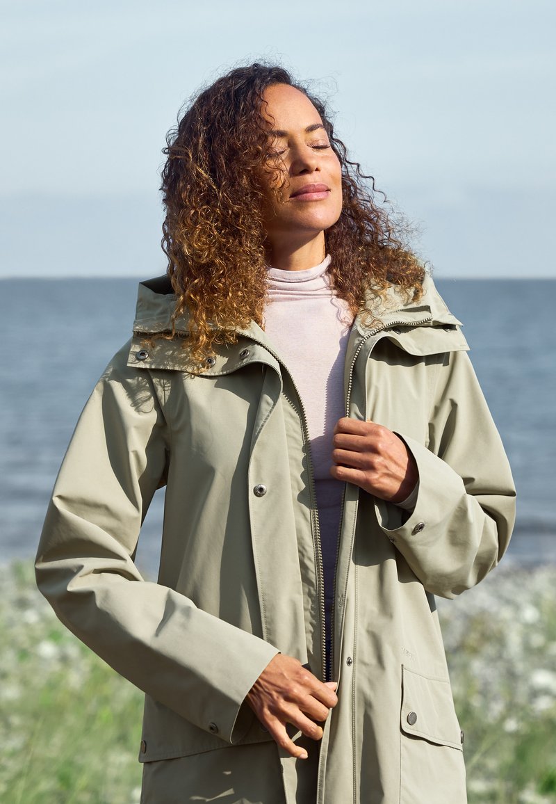 Hellgrüne wasserdichte Jacke mit Kapuze, Reißverschluss und Druckknopf-Taschen. Glatte Textur und lässiges Design, getragen über einem leichten Oberteil.