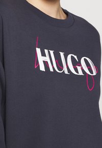 Felpa scura blu navy con collo a giro, caratterizzata da un grande logo "HUGO" in bianco e rosa. Realizzata in un tessuto morbido con una superficie liscia.