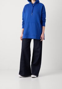 Sudadera de algodón azul con bolsillo frontal, diseño oversize y capucha con cordón, combinada con jeans oscuros de pernera ancha y zapatillas blancas.