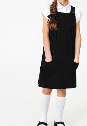 Jeune fille portant une robe noire sans manches avec des poches par-dessus un chemisier blanc à manches courtes, des chaussettes blanches montantes jusqu'aux genoux et des chaussures noires.