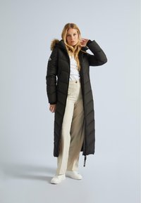 Femeie îmbrăcată cu un palton lung negru matlasat, cu glugă căptușită cu blană, top alb, pantaloni bej cu croi larg și adidași albi, stând în fața unui fundal simplu.