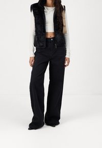 Schwarze Kunstpelz-Weste über einem weißen Langarmoberteil, kombiniert mit hochtaillierten, weiten schwarzen Jeans und spitzen schwarzen Absätzen.