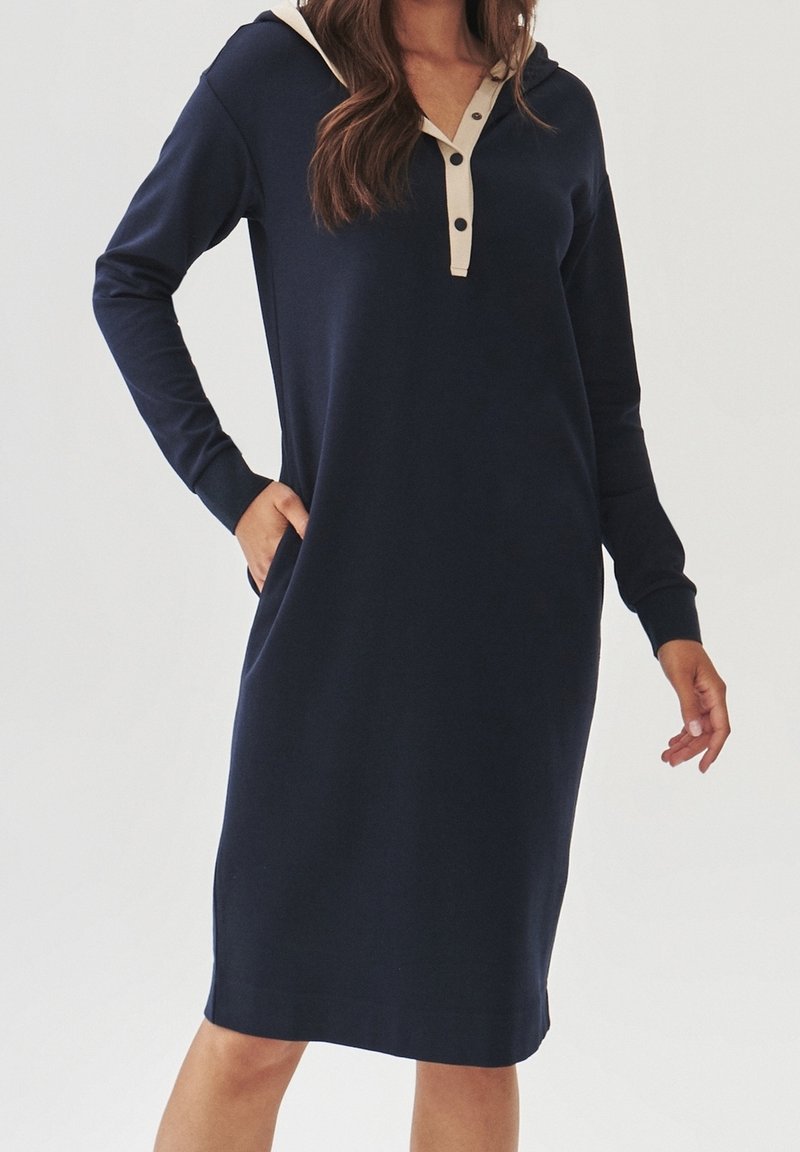 Femme en robe à capuche bleu marine à manches longues avec patte de boutonnage beige, main dans la poche latérale, debout devant un fond uni.