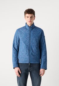 Casaco azul com fecho éclair, apresentando um padrão camuflado texturizado, gola alta e mangas compridas, combinado com jeans escuros. Material leve.