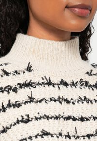 Gros plan sur une personne portant un pull à col roulé crème avec des rayures horizontales noires texturées.