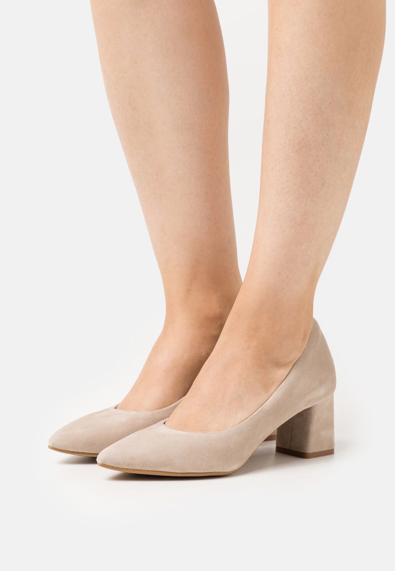 Chaussures pour femmes en daim beige à talon bloc, portées pieds nus, avec des bouts pointus et des talons de hauteur moyenne sur fond blanc.