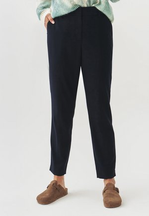 Marineblauwe, op maat gemaakte broek met een slanke pasvorm en een crop-lengte. Gladde stof met strakke lijnen, gecombineerd met bruine suède klompen.