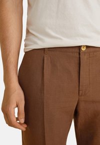 Pantaloni in lino marrone con taglio dritto, dotati di chiusura frontale con bottone e tasche laterali. Il tessuto ha una finitura liscia e texturizzata.
