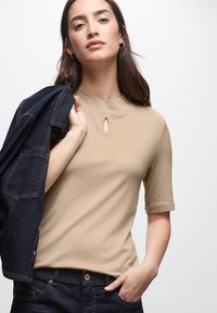 Kvinde iført en beige kortærmet top med nøglehul detalje, som holder en mørk denimjakke over skulderen, med den ene hånd i lommen på sine blå jeans.