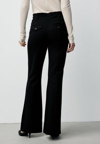 Black flared trousers with a high waist, featuring two back pockets with button closures and a smooth fabric texture.