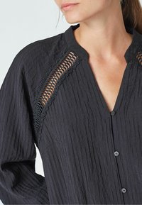 Camicia nera con texture, scollo a V e dettagli in pizzo lungo le spalle, dotata di chiusure a bottoni e maniche lunghe e morbide.
