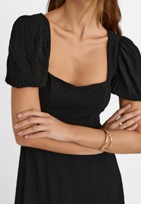 Schwarzes texturiertes Kleid mit Puffärmeln und eckigem Ausschnitt. Ein goldenes Armband schmückt ein Handgelenk, die Arme sind vor der Brust verschränkt.