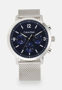 Reloj de malla de acero inoxidable plateado con una esfera azul oscura, marcadores de hora blancos, tres subesferas y una pantalla de fecha. Presenta funciones de cronógrafo.