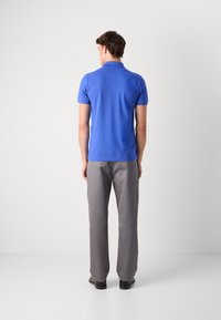 Camisa polo em azul sólido, com mangas curtas e colarinho. Combinada com calças cinza claro e sapatos pretos, vista de trás.