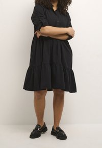 Schwarzes, knielanges Kleid mit durchgehender Knopfleiste, umgeschlagenen Ärmeln und gestuftem Rock. Getragen mit schwarzen Slippern.