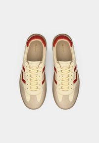 Baskets basses en cuir crème avec des accents rouges et du daim beige. Elles présentent des lacets plats, un bout arrondi et un logo sur la languette.