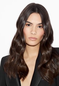 Vrouw met lang, golvend, donkerbruin haar, lichte make-up en een neutrale uitdrukking, gekleed in een zwarte blazer tegen een witte achtergrond.