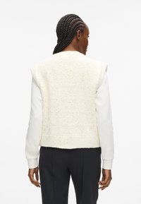 Gaišs krēms vestes ar teksturētu, punktveida adījumu. Bez piedurknēm, ribota apakšmala un kontrastējošas baltas garās piedurknes.