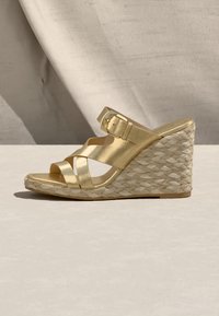 Sandale compensée dorée à brides, talon style espadrille tissé et bride ajustable avec boucle, sur un fond texturé neutre.