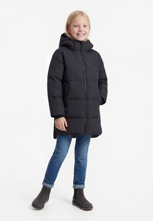 Bambino sorridente con giacca nera imbottita lunga con cappuccio, jeans blu risvoltati e stivaletti scuri, in piedi davanti a uno sfondo bianco.