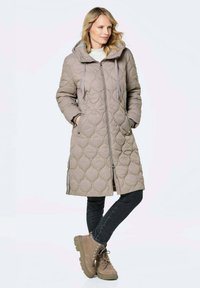 Gepolsterte, hüftlange, beige Parka mit Kapuze, Reißverschluss und seitlichen Taschen. Kombiniert mit dunklen Jeans und hellbraunen Stiefeletten.