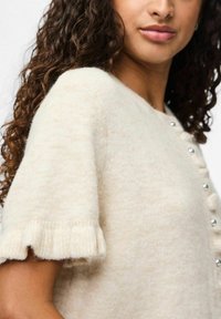 Krémový svetr s měkkým, texturovaným povrchem. Má kulatý výstřih, krátké rukávy s volánky a stříbrné knoflíkové detaily.