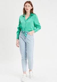 Chemise en satin vert à boutons, associée à un jean taille haute bleu clair, avec une ceinture à motif floral. Portée avec des baskets blanches.