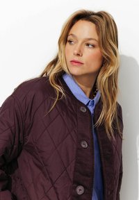 Femme blonde regardant à gauche, portant une veste matelassée violette foncée avec de grands boutons sur une chemise col bleue, sur fond blanc uni.