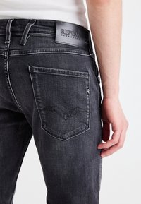 Personne portant un jean noir Replay avec des coutures visibles sur la poche arrière et un patch en cuir de la marque sur la ceinture.