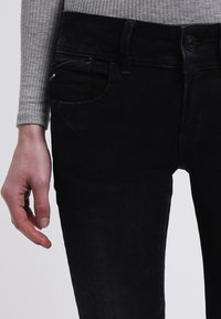 Jeans de mezclilla negra con un corte ajustado, que presentan un bolsillo frontal, trabillas para el cinturón y una superficie texturizada. Acompañados de una camiseta de manga larga acanalada en gris.