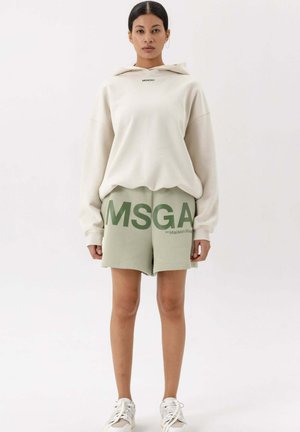Overmaat crèmekleurige hoodie met logo aan de voorkant, gecombineerd met lichtgroene shorts met grote tekst en een op maat gemaakte pasvorm. Sneakers maken de look compleet.