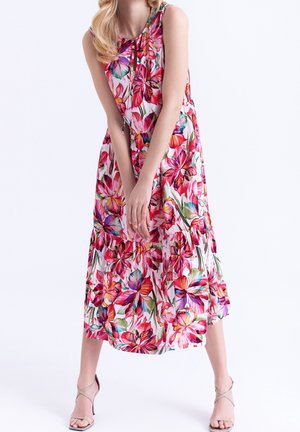 Vrouw draagt een mouwloze witte midi-jurk met grote kleurrijke bloemenprint en beige sandalen met hoge hakken, staat met handen gevouwen.