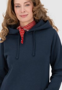 Felpa blu navy con tasca frontale, cappuccio con stringa e texture morbida; abbinata a una sciarpa rossa a pois al collo.
