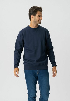 Mand iført en mørkeblå langærmet sweatshirt og blå jeans, stående og kiggende til siden mod en ensfarvet baggrund.