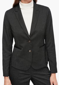 Czarny żakiet o teksturowanej fakturze i dopasowanym kroju, z dwoma guzikami i bocznymi kieszeniami. Nosi się go na białym golfie. Delikatne szczegóły w kratkę.