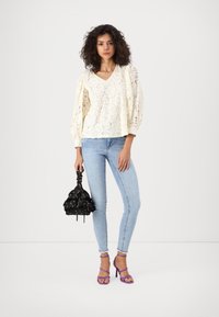 Kvinne med krøllete hår iført en hvit bluse i blonder, lyseblå stramme jeans, lilla hæl sandaler, og holder en liten svart blomstrete håndveske.