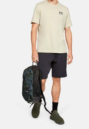 Mann trägt beiges Under Armour T-Shirt, schwarze Shorts, grüne Sneakers, hält einen schwarzen und camouflagefarbenen Under Armour Rucksack am obersten Griff.