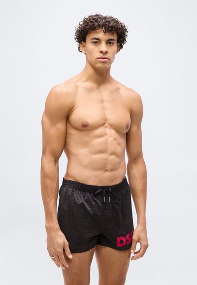 Jeune homme aux cheveux bouclés portant un short de bain noir avec le logo rose "DSQ", debout devant un fond clair uni.