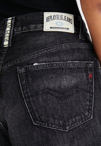 Čierne denimové džínsy s modro-bielou nálepkou "Replay Blue Jeans". Majú zadnú vrecko s červeným prešívaním a textúrovaný povrch.