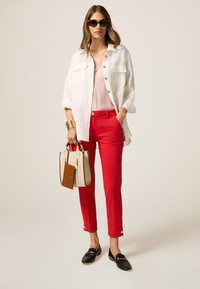 Camicia bianca in lino con bottoni, canotta rosa, pantaloni rossi su misura, mocassini neri e una borsa beige con accenti marroni. Gli occhiali da sole completano il look.
