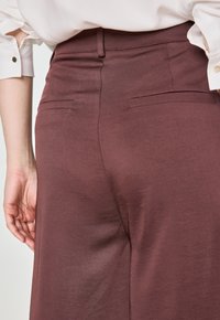 Pantaloni bordeaux con una trama morbida, dotati di una vita standard e due tasche posteriori, parzialmente visibili con una mano in vista.