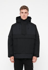 Casaco pullover preto com capuz, apresentando texturas contrastantes, cordões ajustáveis e múltiplos bolsos nas mangas e à frente.