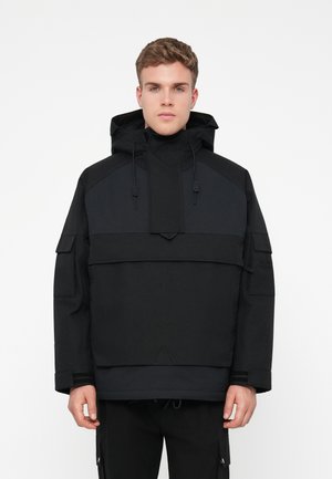 Zwarte pulloverjas met capuchon, voorzien van contrasterende texturen, verstelbare trekkoorden en meerdere zakken op de mouwen en aan de voorkant.