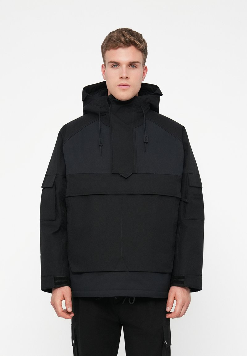 Casaco pullover preto com capuz, apresentando texturas contrastantes, cordões ajustáveis e múltiplos bolsos nas mangas e à frente.