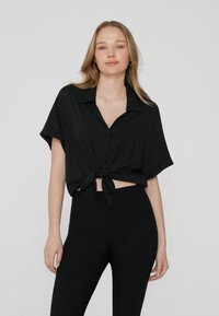 Schwarze Bluse mit kurzen Ärmeln, Kragen und vorderem Knoten-Detail, kombiniert mit hoch taillierten schwarzen Leggings. Glattes Material und lockere Passform.